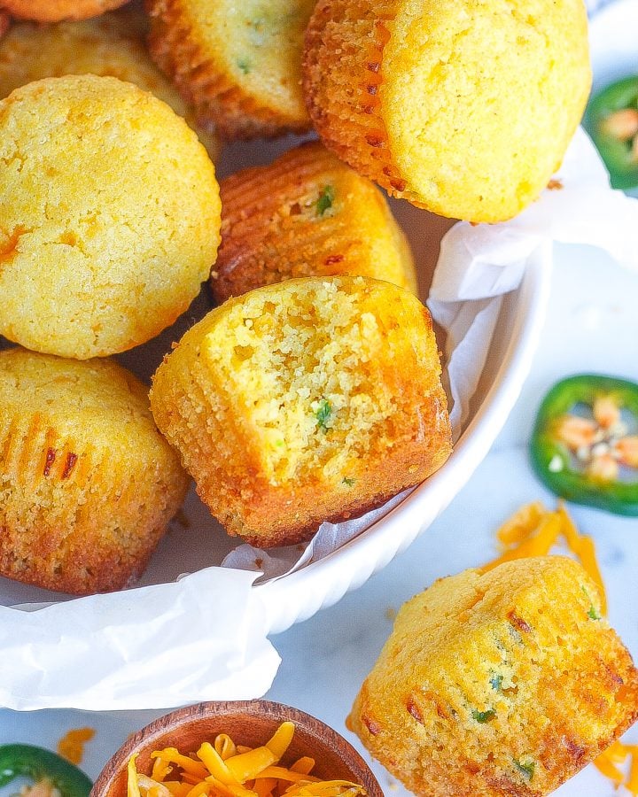 one white bowl of jalapeño cheddar cornbread muffins with one bite taken out of one muffin