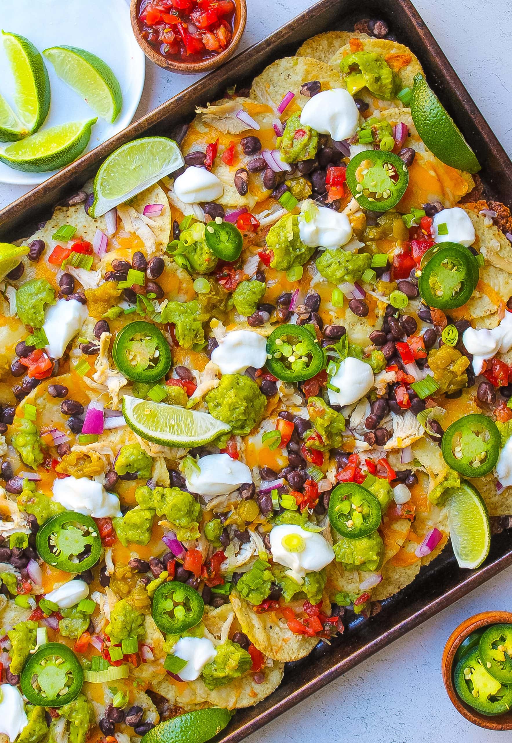 one pan filled with baked chicken nachos with lime, black beans, cheese, sour cream, avocado, pico de gallo, red onion, jalapeńo