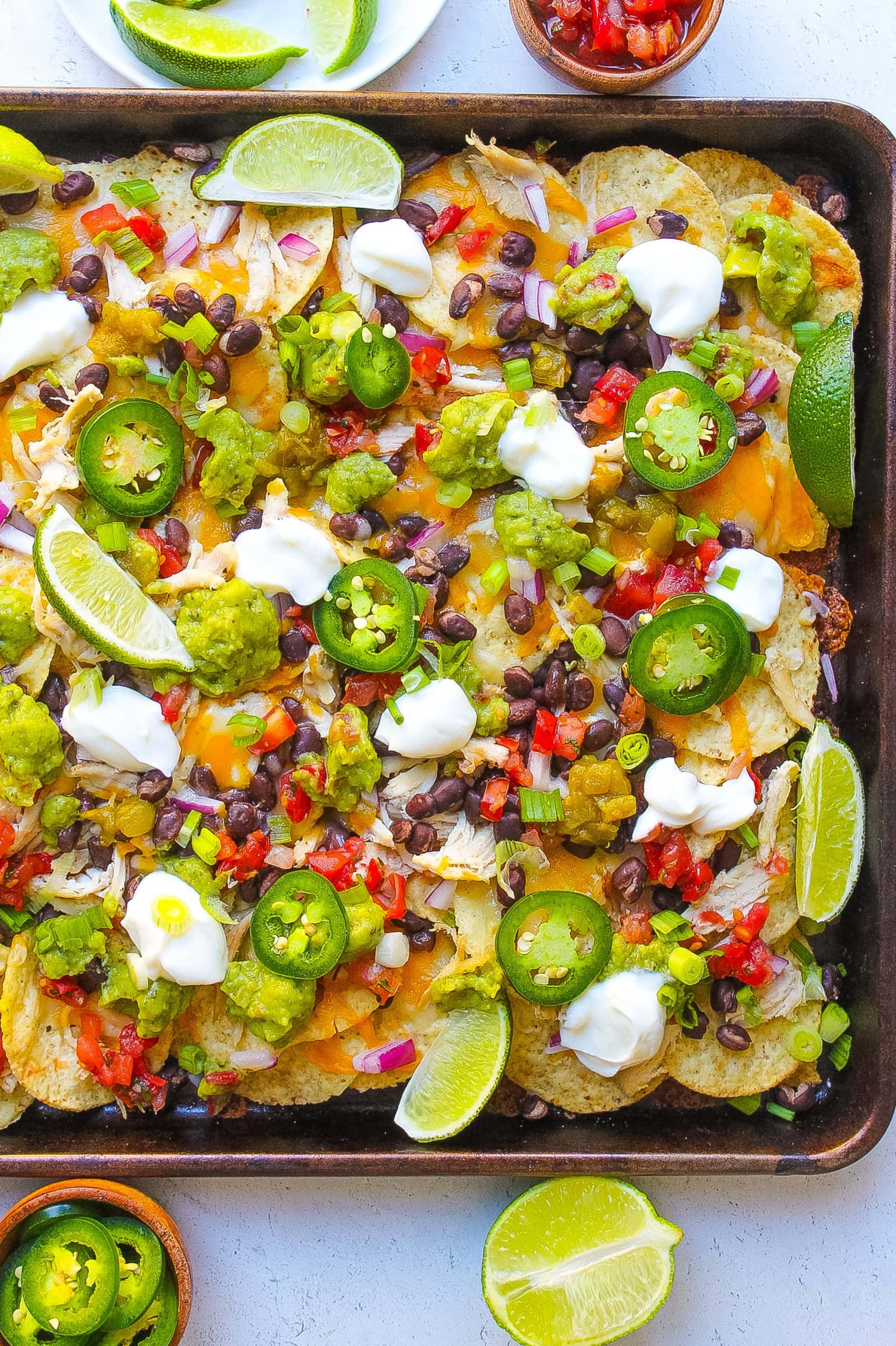one pan filled with baked chicken nachos and topped with sour cream, lime, jalapeńo, cheese