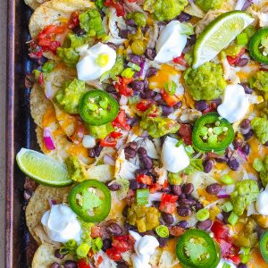 top view of baked chicken nachos with jalapeńo, sour cream, lime, cheese, red onion, black beans, guacamole