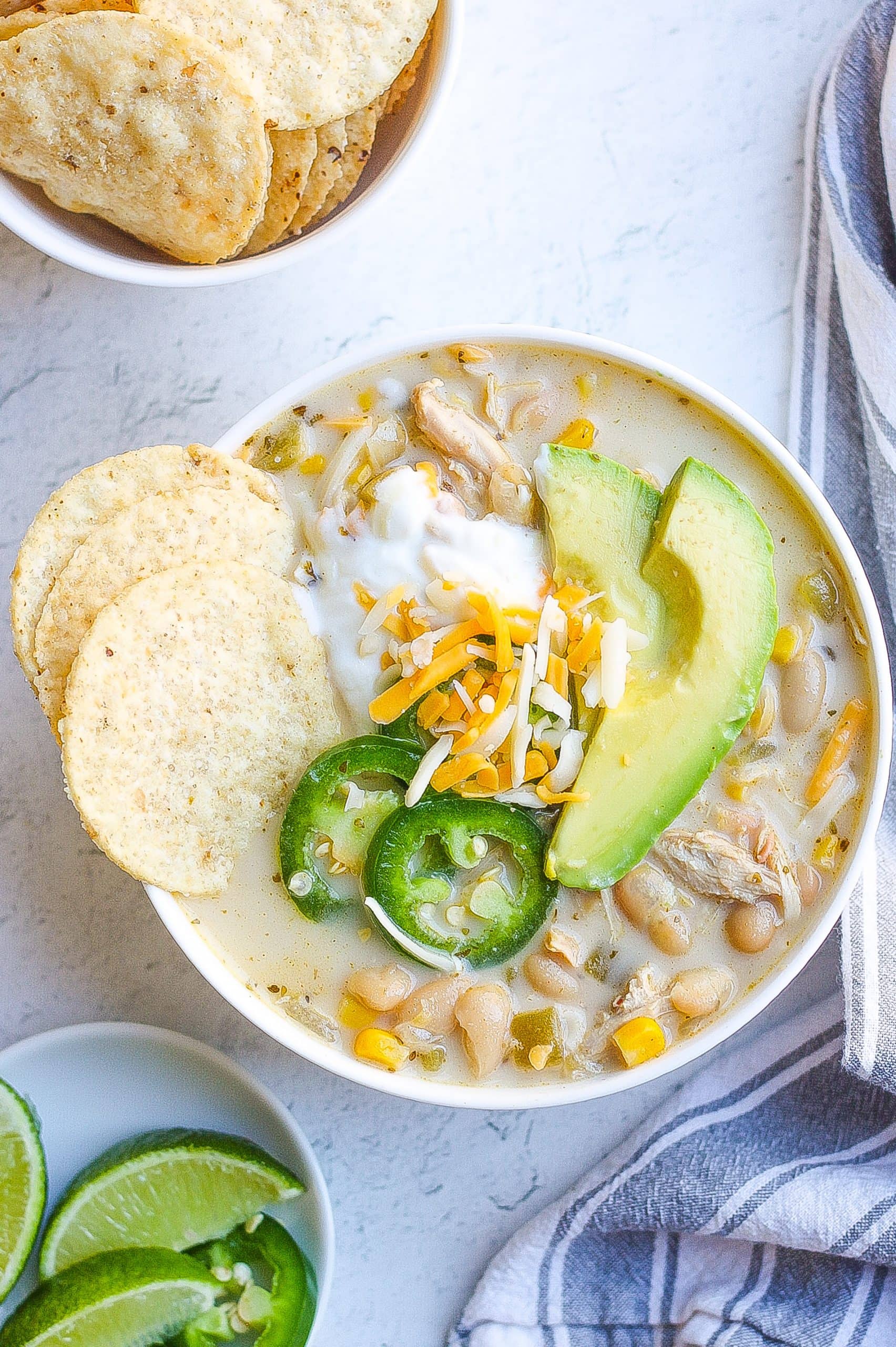 Bowl of white chicken chili topped with tortilla chips, sour cream, shredded cheese, and slice jalapeno and avocado. 