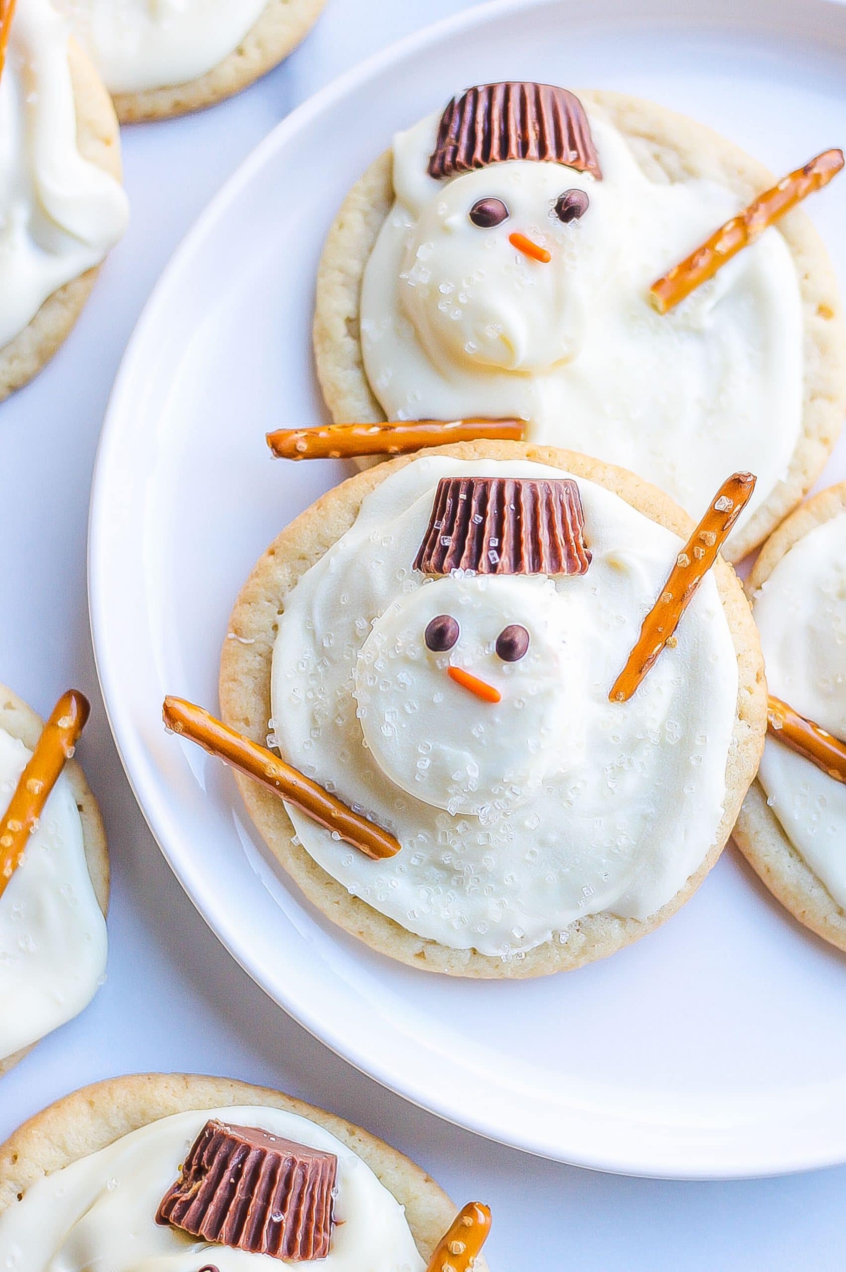 snowman cookies on white plate with pretzels, marshmallows, white candy, chocolate chips, orange jimmies, and Reese's hats