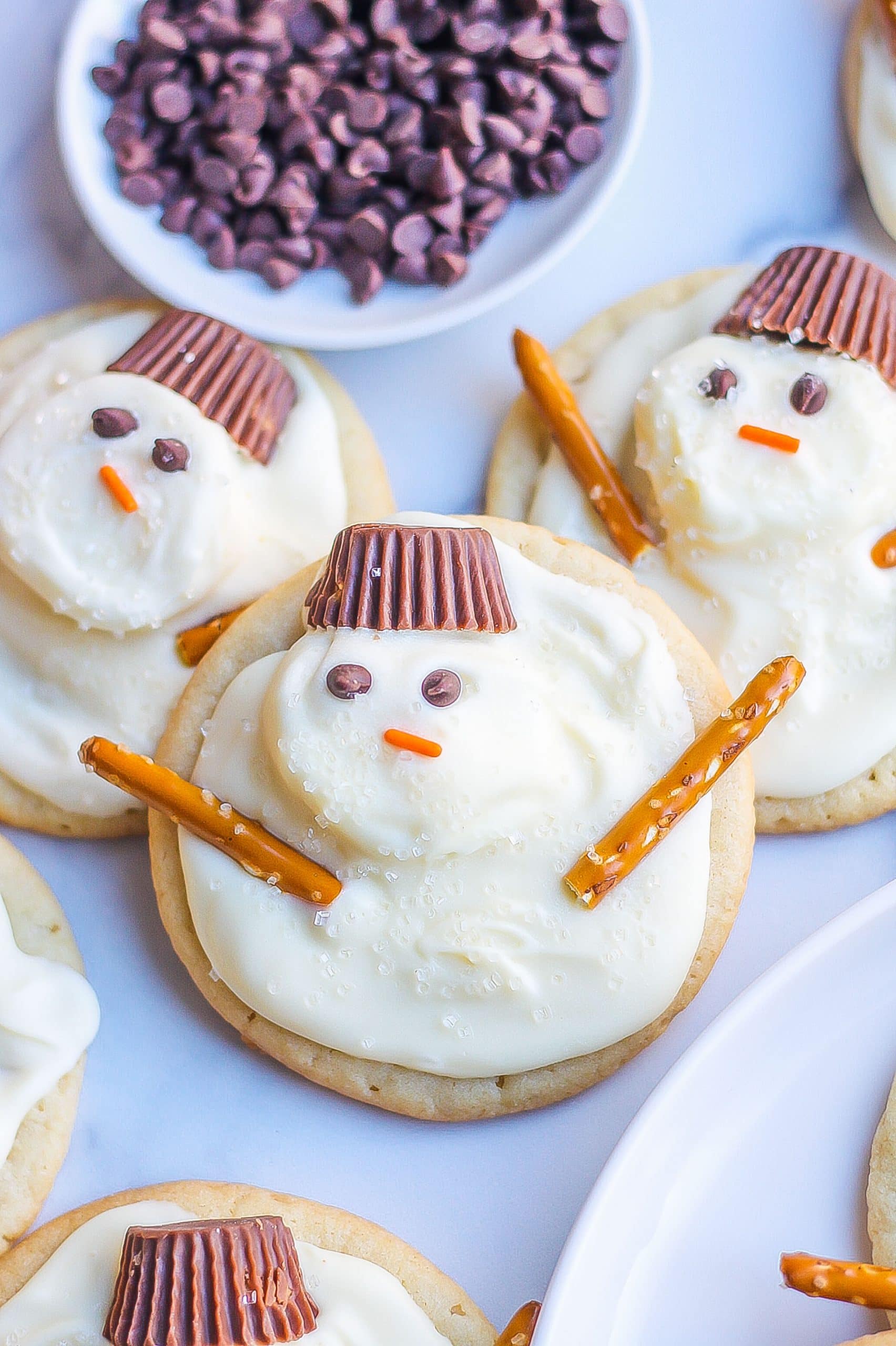 three snowman cookies with chocolate chips, marshmallows, Reese's hats, and pretzel stick arms