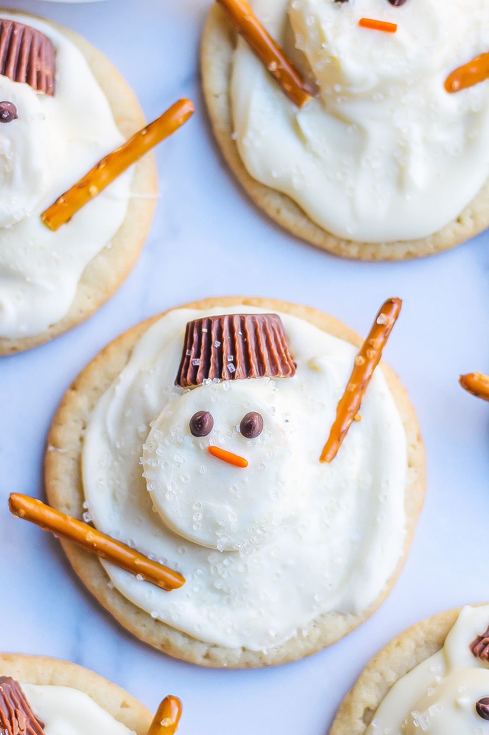 top view of snowman cookies with pretzel arms, eyes, Reese's hats, marshmallows, and sparkling sugar