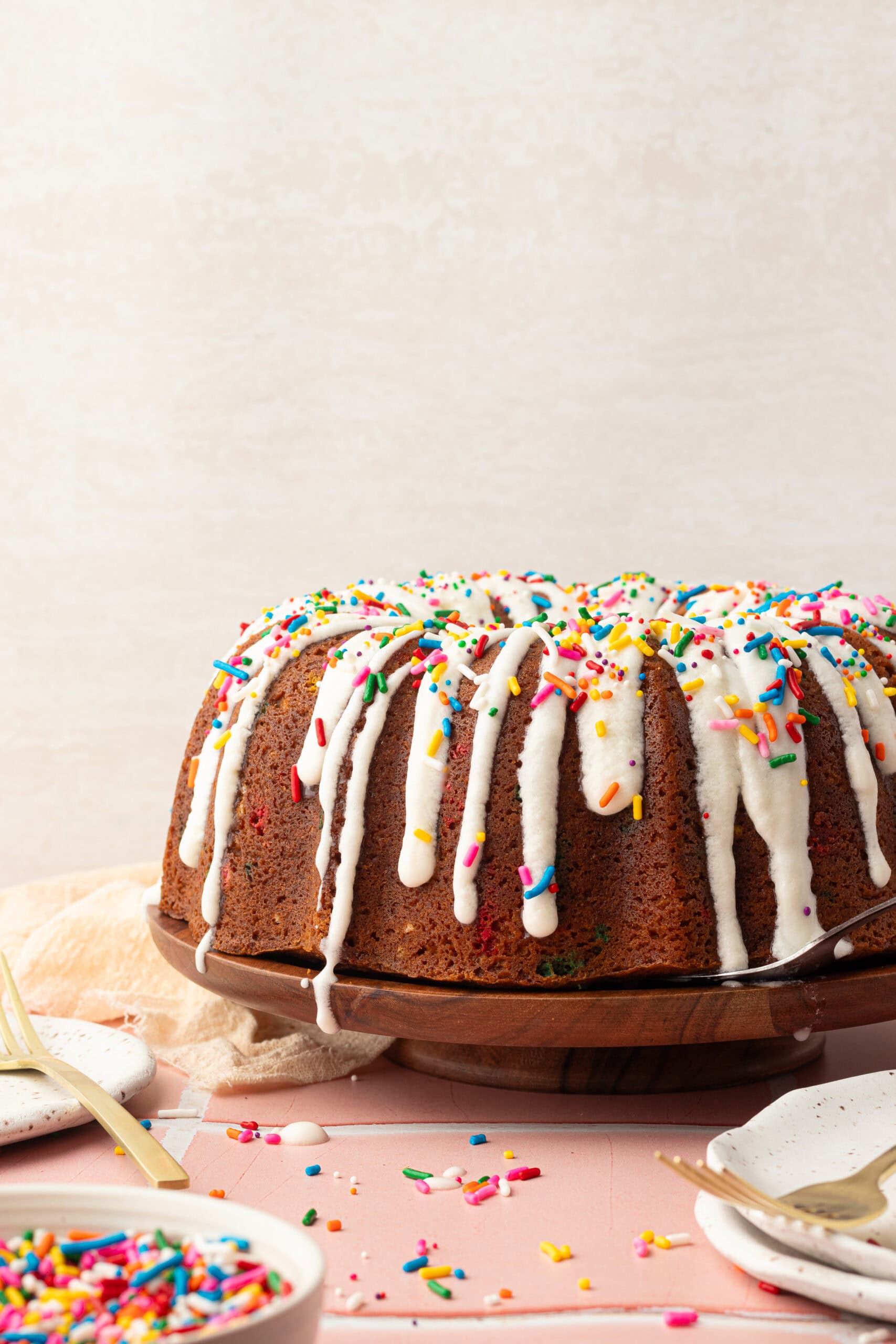 A funfetti bundt cake with frosting dripping down the sides and rainbow sprinkles on top.