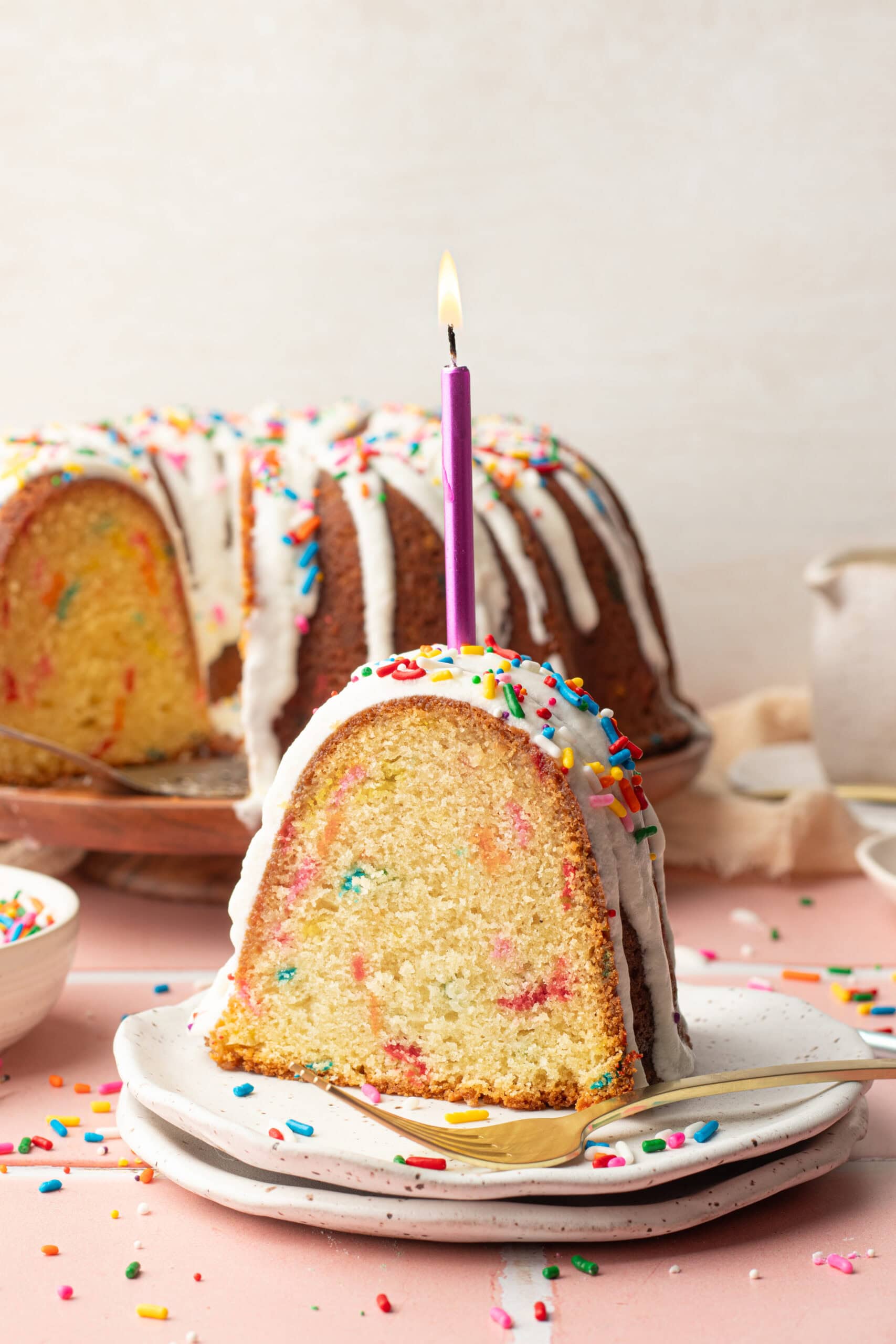 Slice of Funfetti Bundt Cake on a small plate with lots of rainbow sprinkles on top and a lite birthday candle.