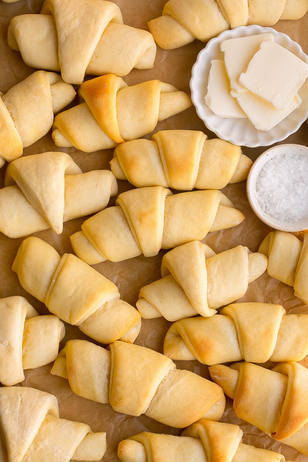 A couple dozen soft homemade crescent rolls on parchment paper with a side of butter and flaky sea salt.