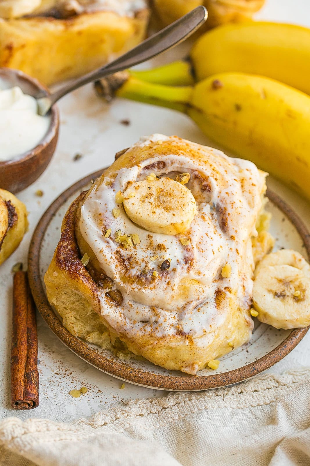 Close-up of a gooey Banana Bread Cinnamon Roll on a plate topped with cream cheese frosting and banana slices.
