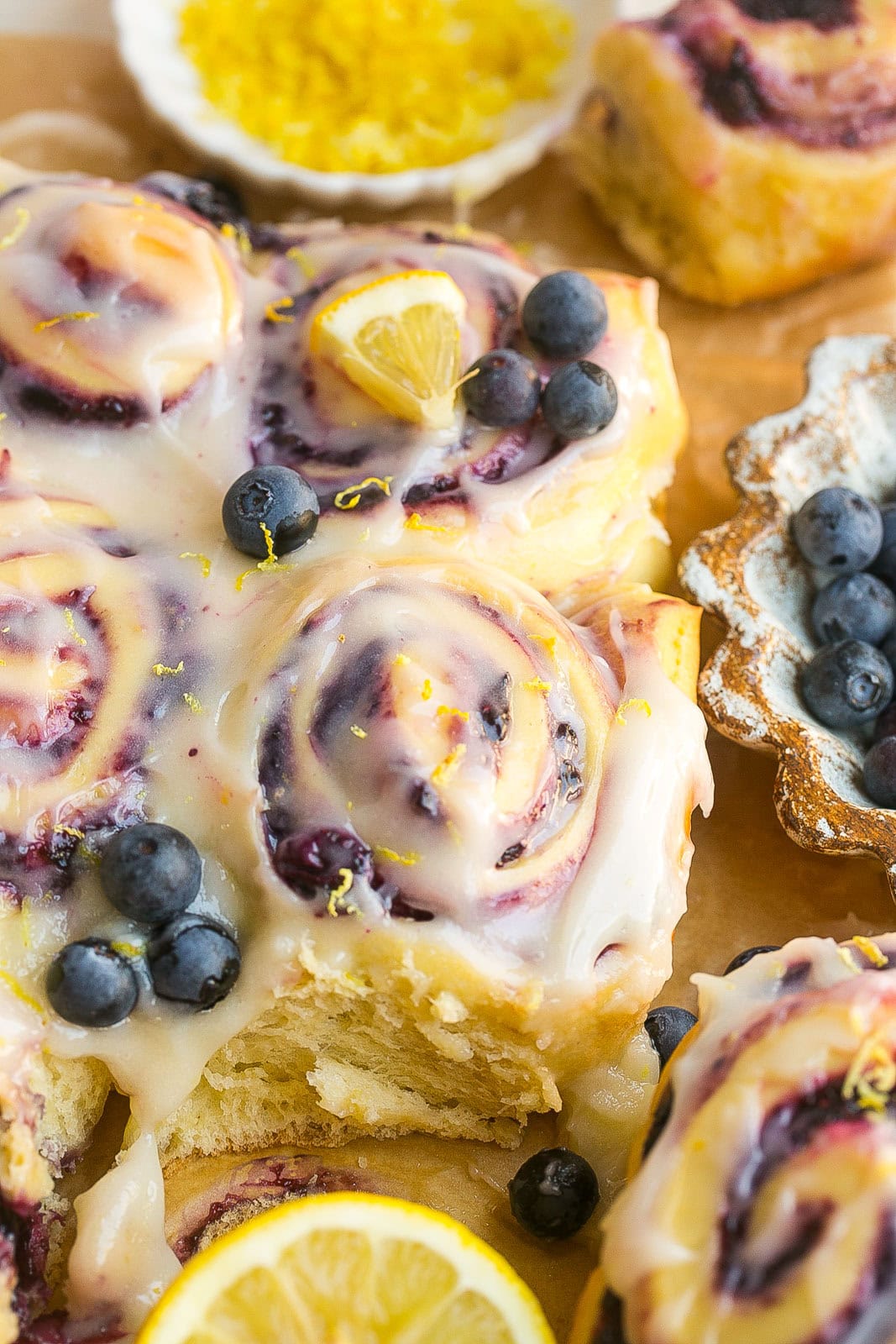Fluffy lemon sweet rolls filled with juicy blueberries and topped with lemon cream cheese icing on parchment paper.