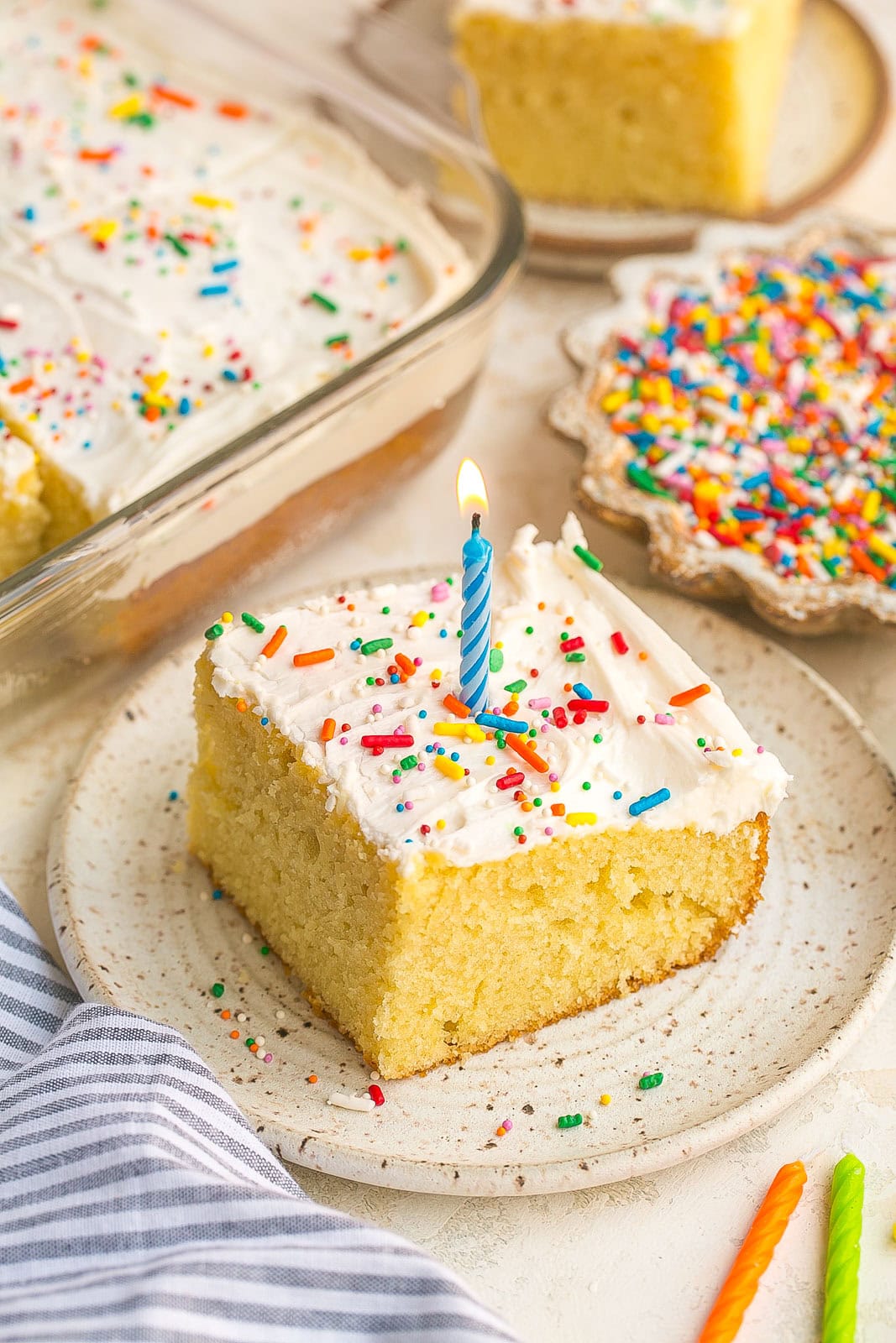 Slice of moist Vanilla Sheet Cake with creamy vanilla buttercream frosting on a dessert plate (with blue candle).